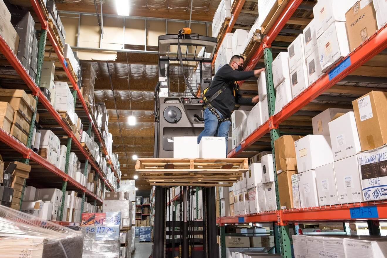 A lifted forklift with a warehouse employee picking out specific cases of wine.